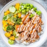 Diced teriyaki salmon with mango salad, edamame, and rice drizzled with spicy mayo in a grey bowl on a grey background.