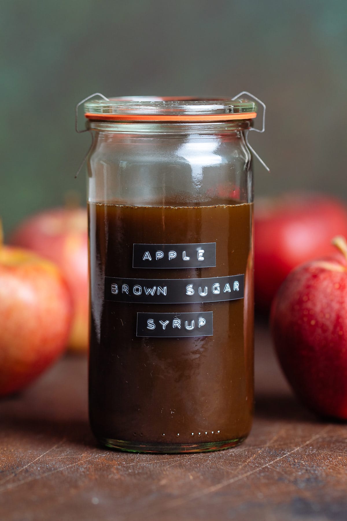 Brown syrup in a glass jar with a label on it that says Apple Brown Sugar Syrup with apples around it on a dark wooden background.