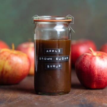 Brown syrup in a glass jar with a label on it that says Apple Brown Sugar Syrup with apples around it on a dark wooden background.
