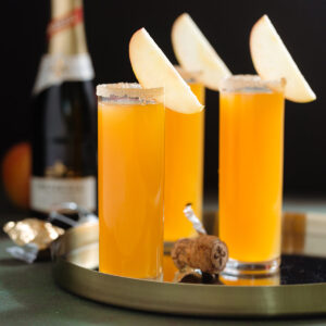 Three tall stemless flute glasses with bright orange apple cider mimosa garnished with a slice of apple and a sugar rim on black background.