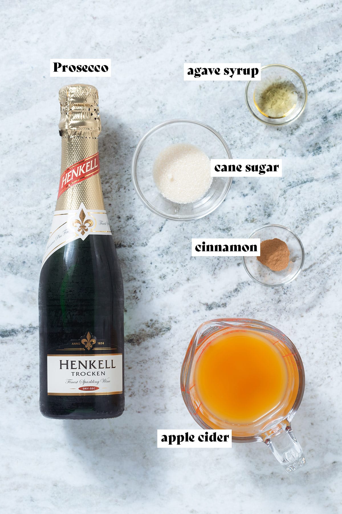 A bottle of prosecco, apple cider, and other mimosa ingredients laid out on a grey stone background with text overlay.