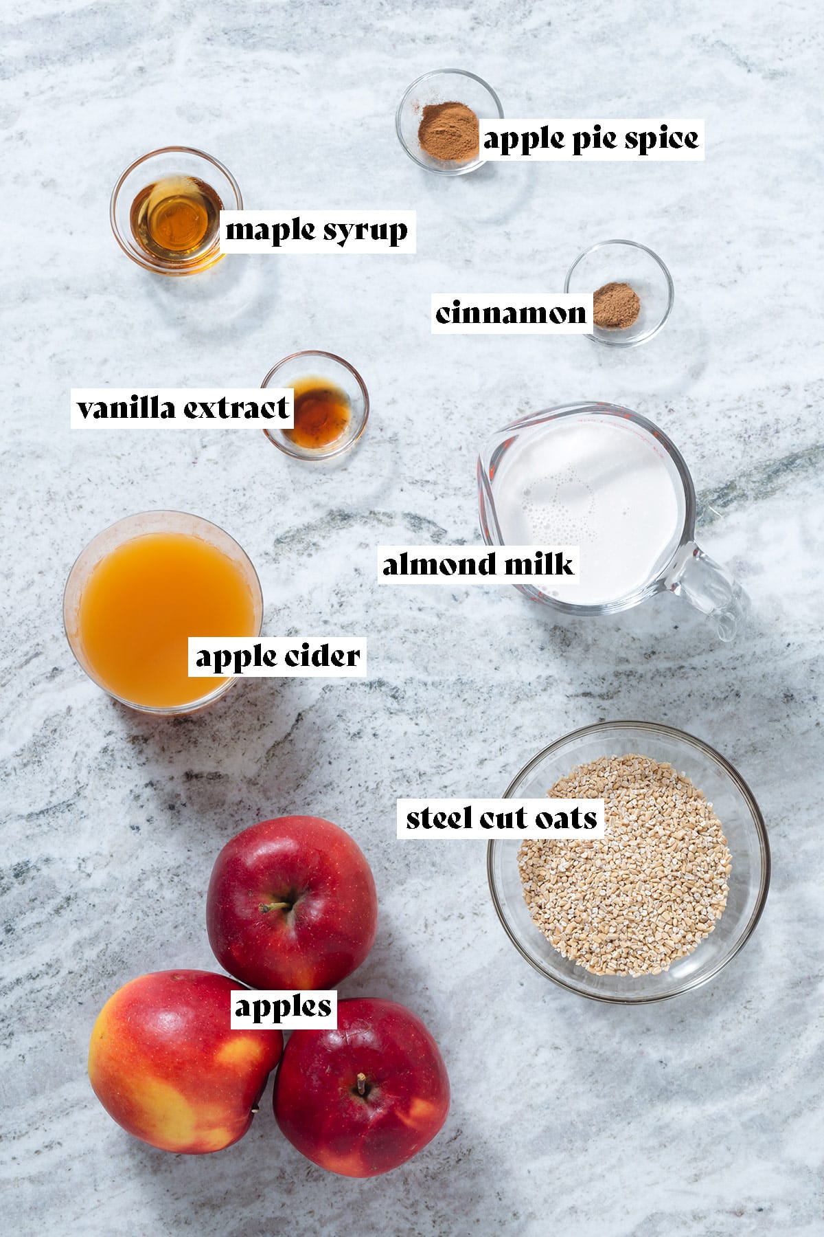 Apples, apple cider, steel cut oats, almond milk, and other measured out ingredients in glass bowls on a grey stone background with text overlay.