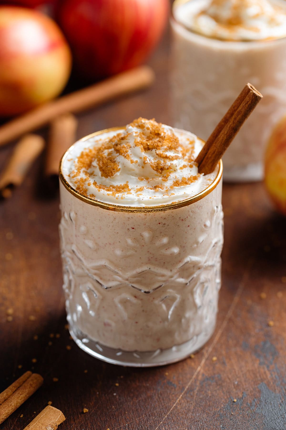 Apple pie smoothie with whipped cream and spiced sugar on top in a short glass with a gold rim garnished with a stick of cinnamon with apples in the background.