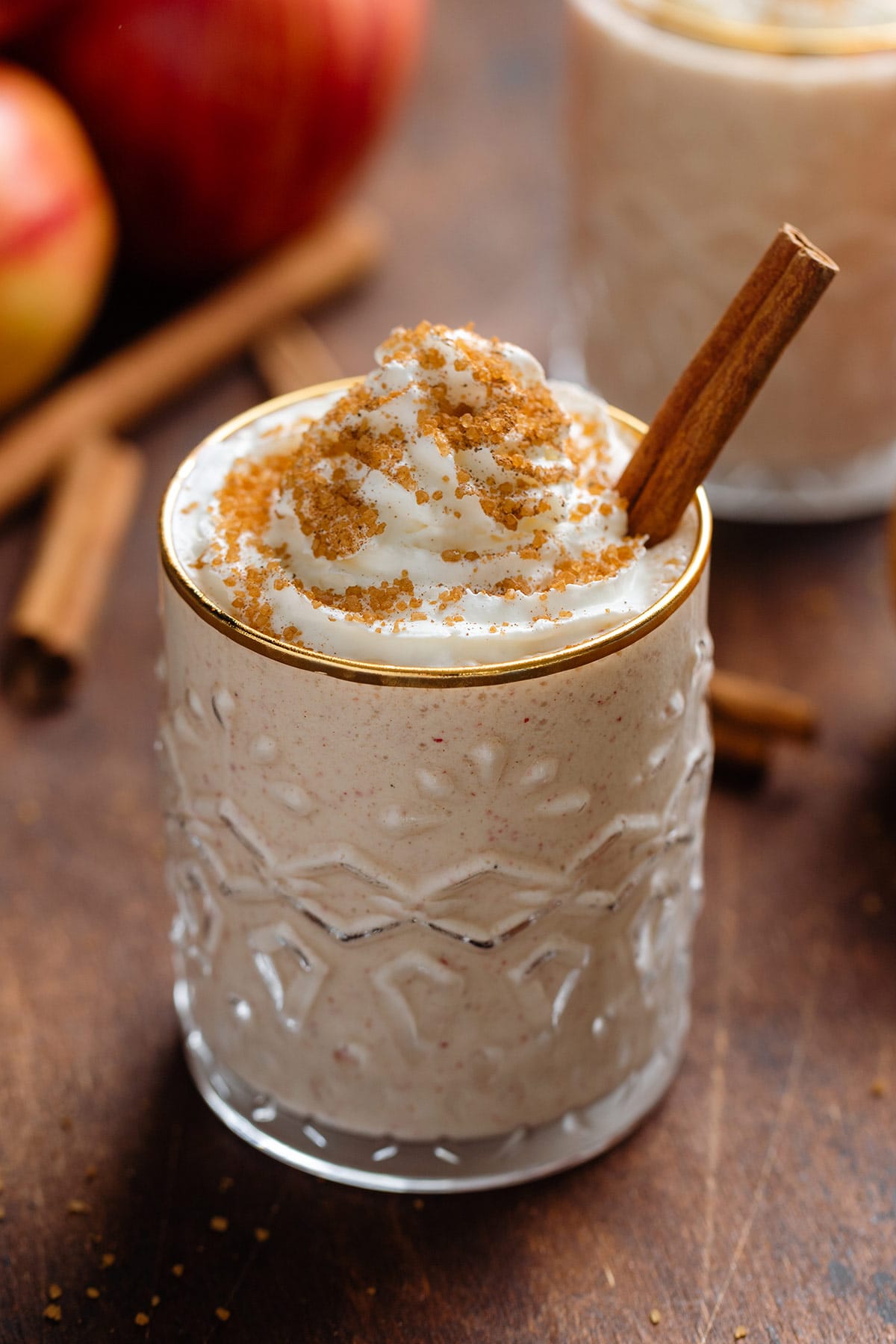 Apple pie smoothie with whipped cream and spiced sugar on top in a short glass with a gold rim garnished with a stick of cinnamon.