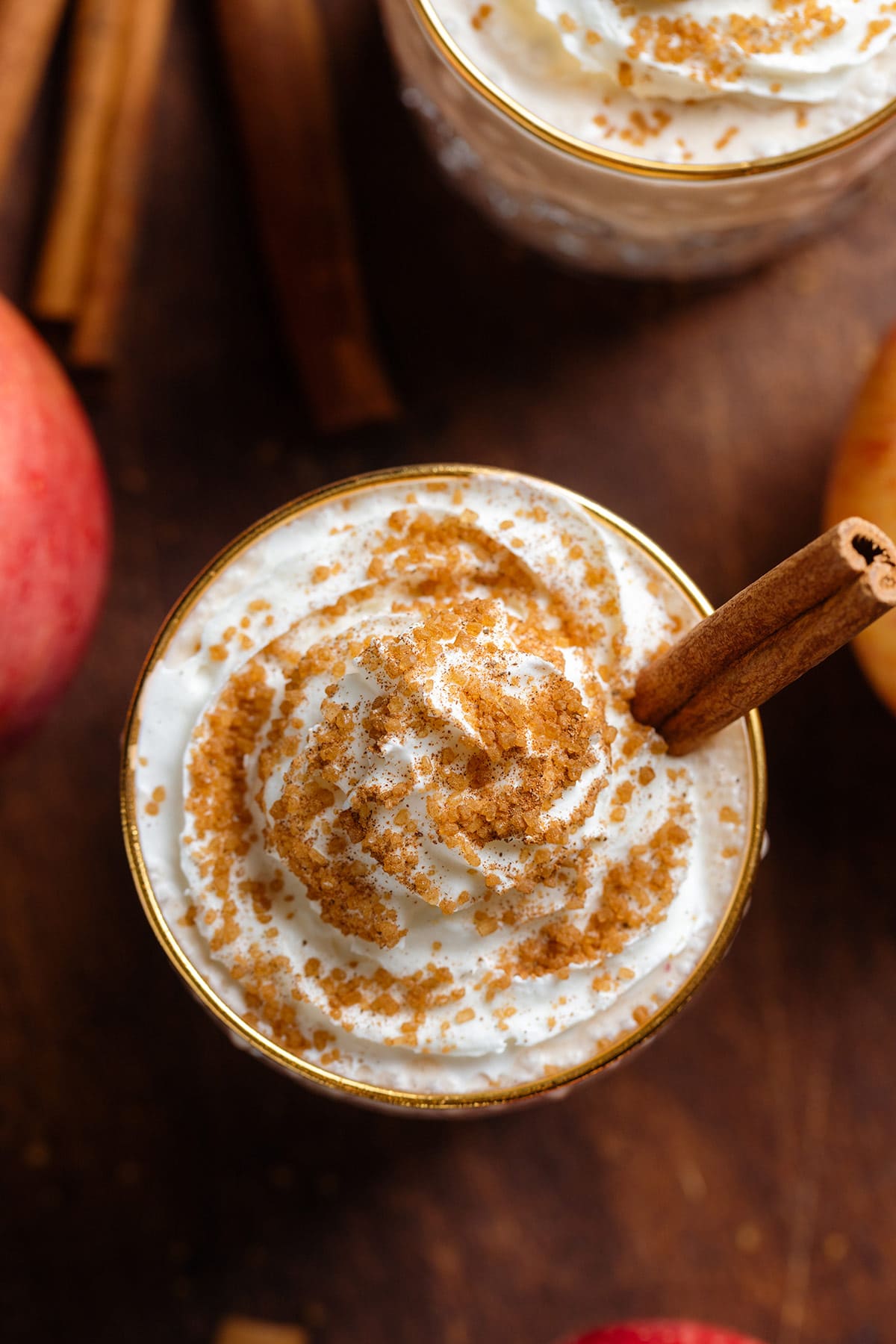 Apple pie smoothie with whipped cream and spiced sugar on top in a short glass with a gold rim garnished with a stick of cinnamon.