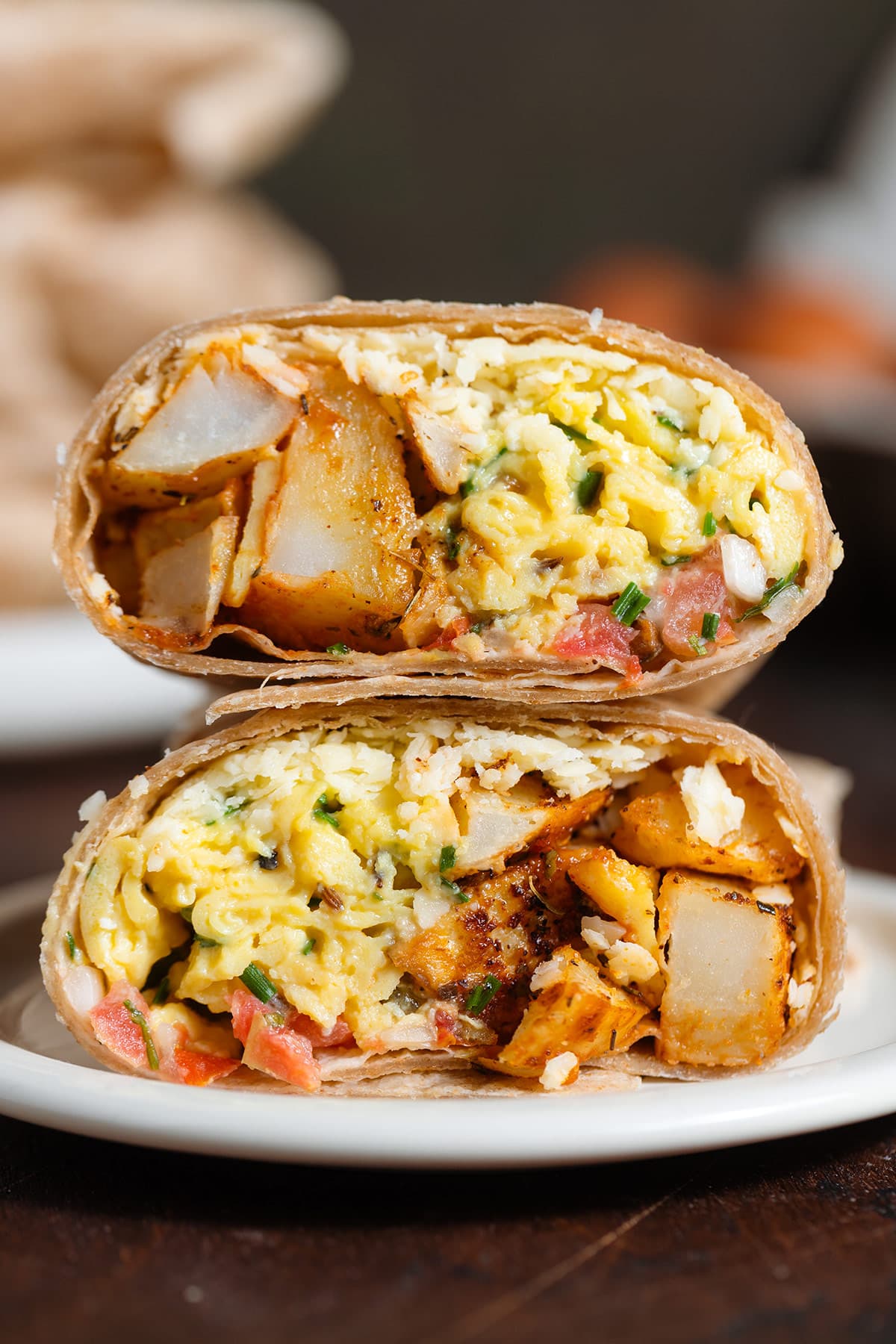 A breakfast burrito cut in half on a small white plate on a wooden background with more burritos and eggs in the background.