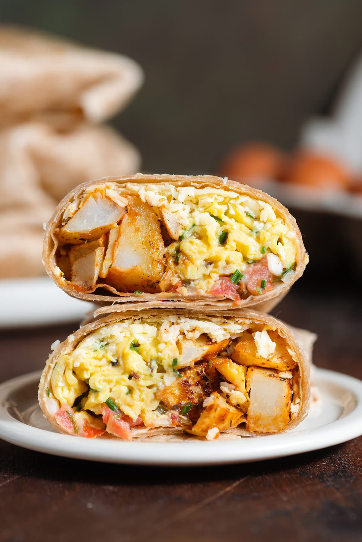 A breakfast burrito cut in half on a small white plate on a wooden background with more burritos and eggs in the background.
