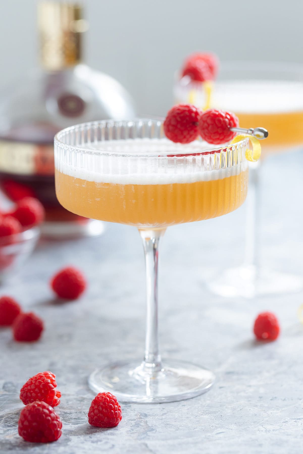 A martini glass with light orange colored cocktail with foam on top garnished with two raspberries on a cocktail pick and a lemon twist with more raspberries around and another cocktail in the background.
