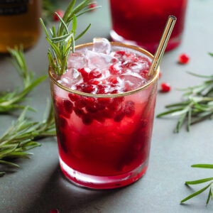 Bright red pomegranate mocktail in a short glass with a gold rim and a gold metal straw garnished with pomegranate arils and fresh rosemary on a green background.