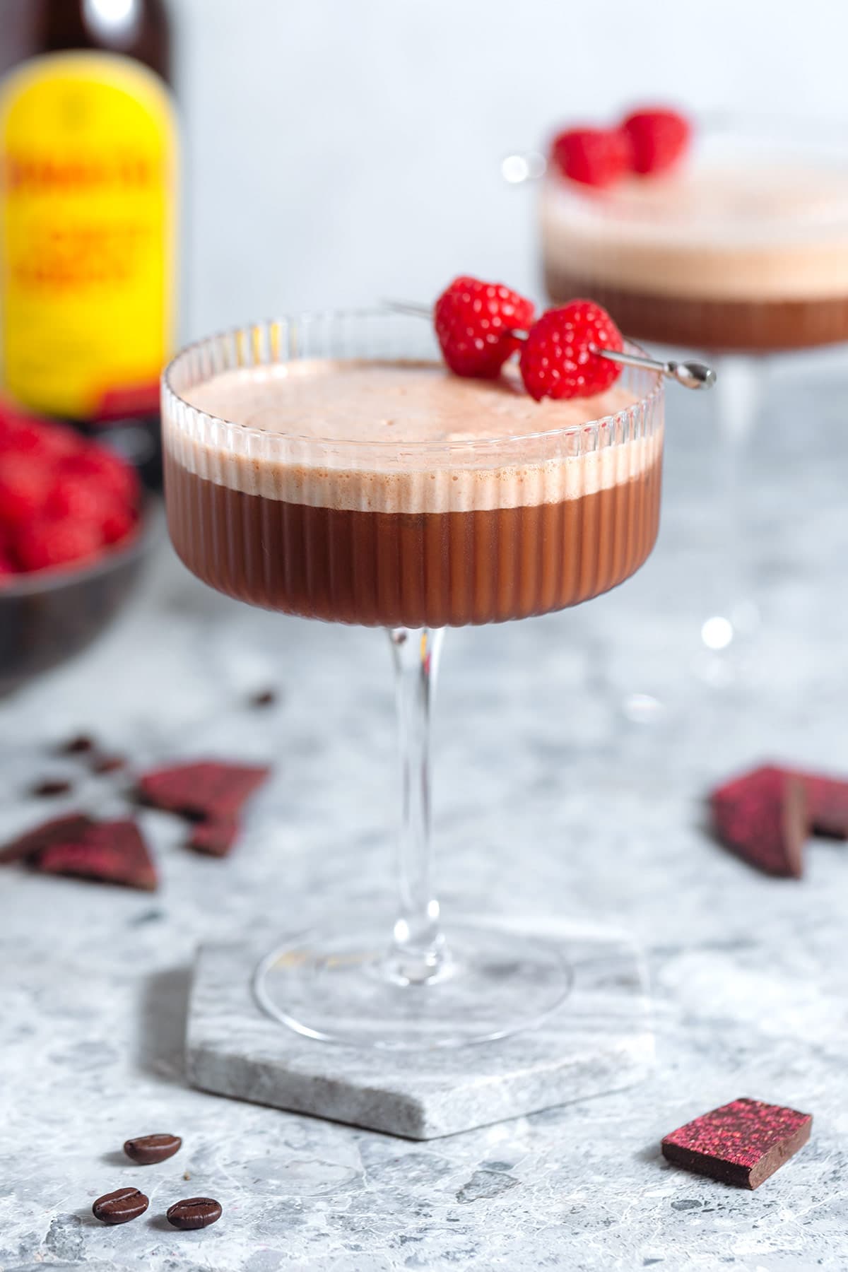 A chocolate espresso martini with rich foam on top in a coupe glass garnished with three coffee beans and a cocktail pick with two raspberries on a grey background with another cocktail in the background.