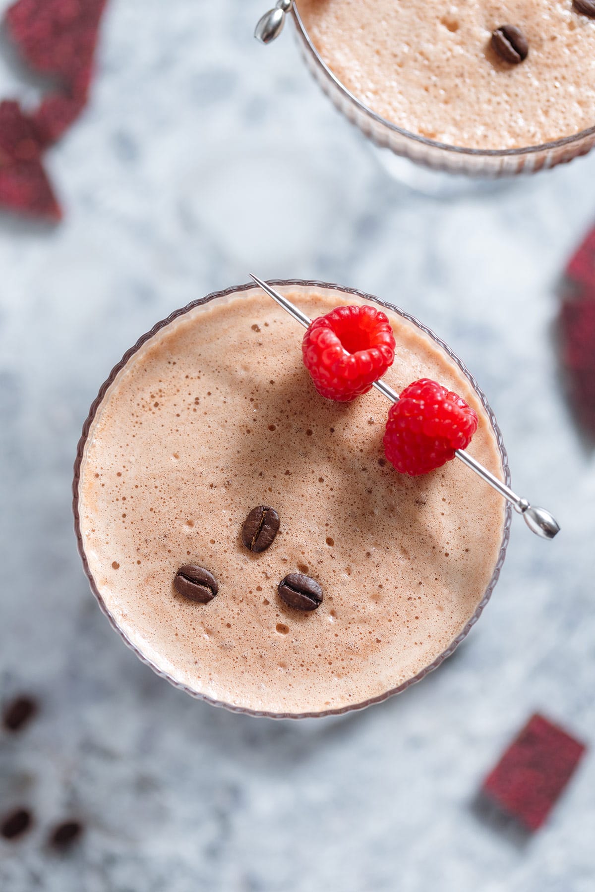 A chocolate espresso martini with rich foam on top in a coupe glass garnished with three coffee beans and a cocktail pick with two raspberries on a grey background shot from above.