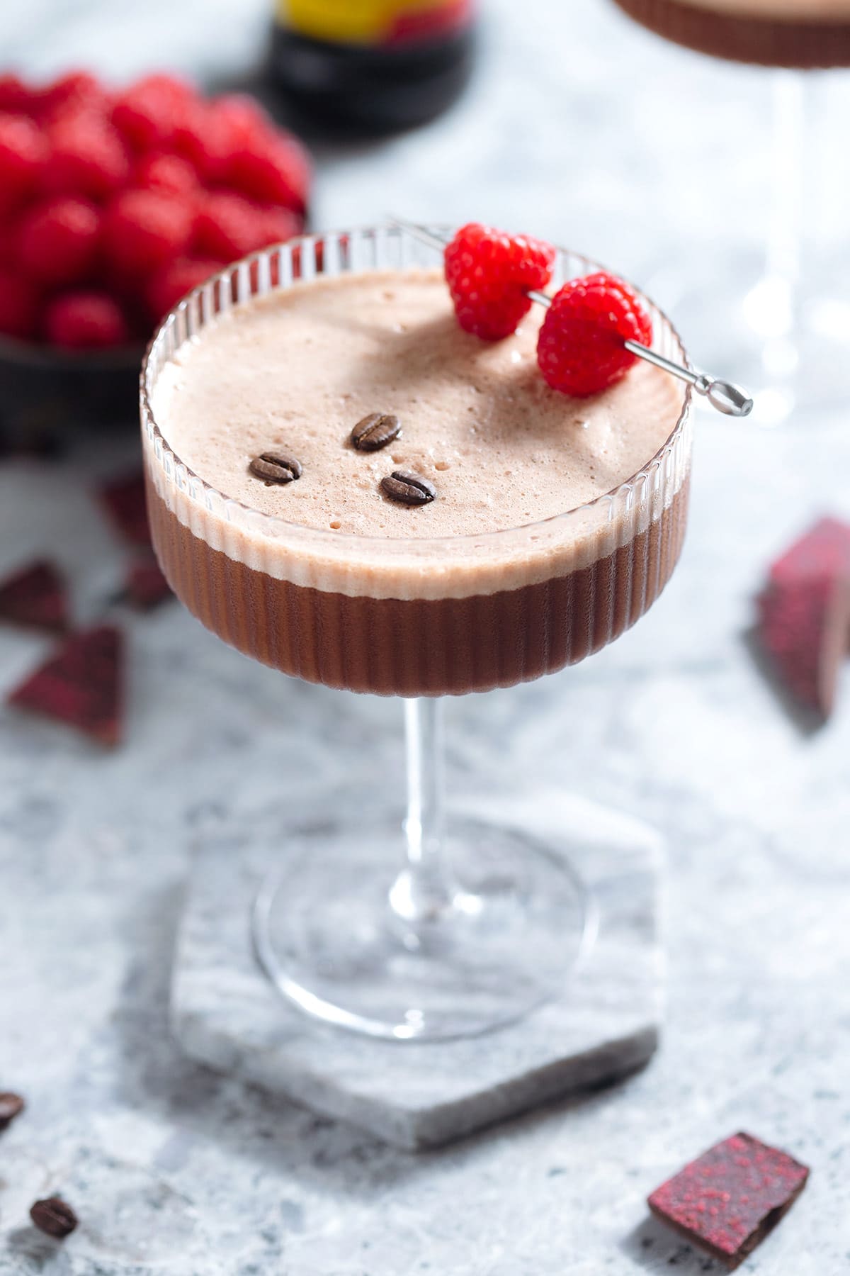 A chocolate espresso martini with rich foam on top in a coupe glass garnished with three coffee beans and a cocktail pick with two raspberries on a grey background.