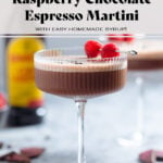 A chocolate espresso martini with rich foam on top in a coupe glass garnished with three coffee beans and a cocktail pick with two raspberries on a grey background.