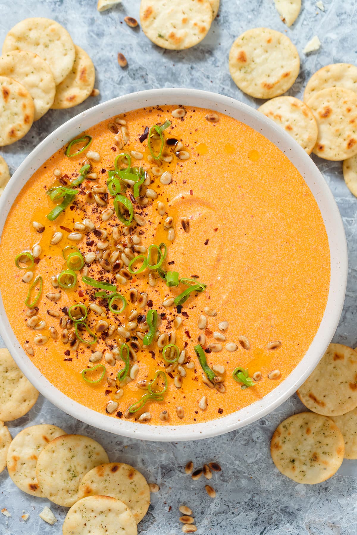 Bright orange dip in a beige bowl garnished with toasted pine nuts, spring onion, and ground sumac, with round crackers around the bowl.