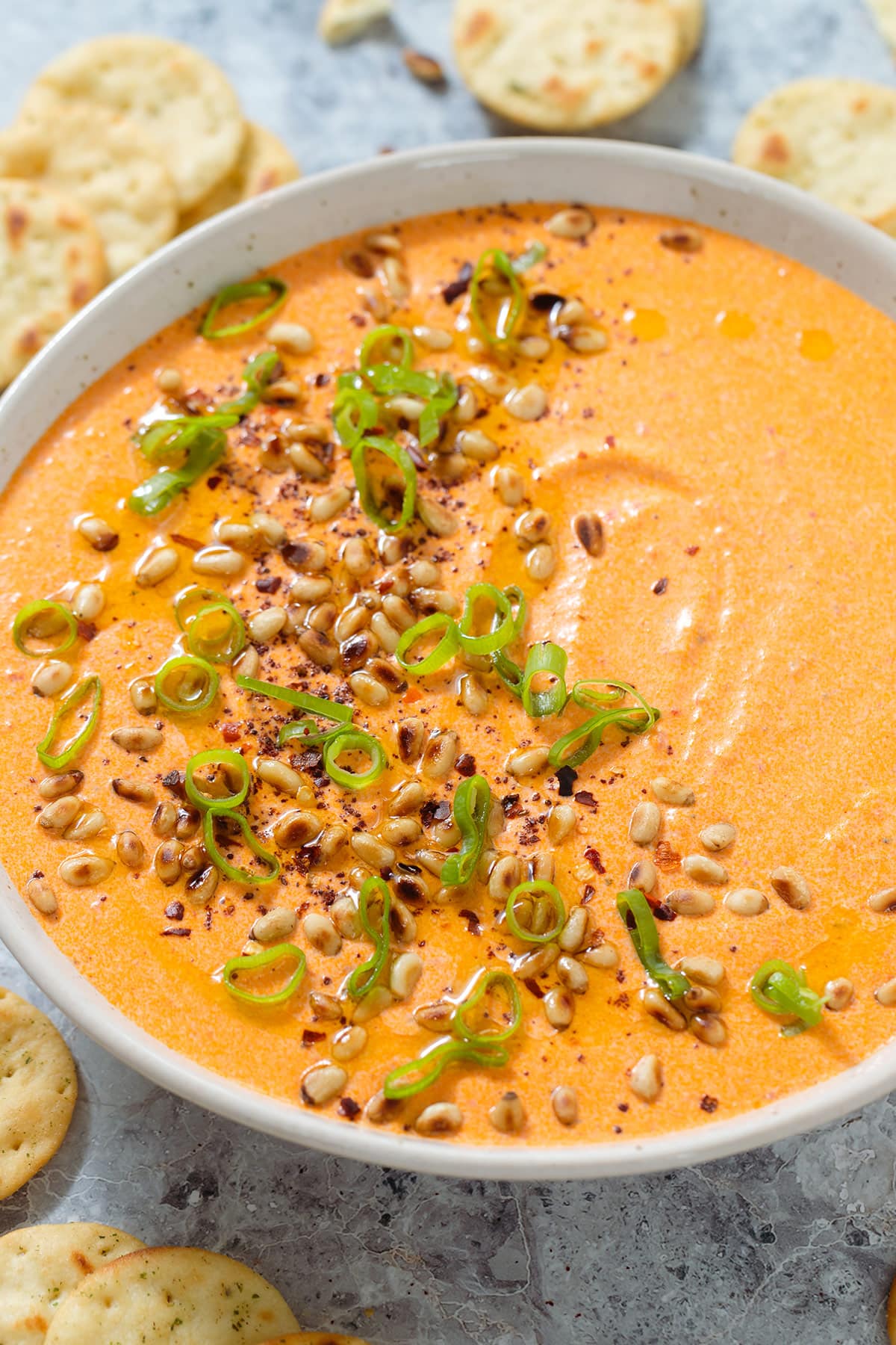 Bright orange dip in a beige bowl garnished with toasted pine nuts, spring onion, and ground sumac, with round crackers around the bowl.