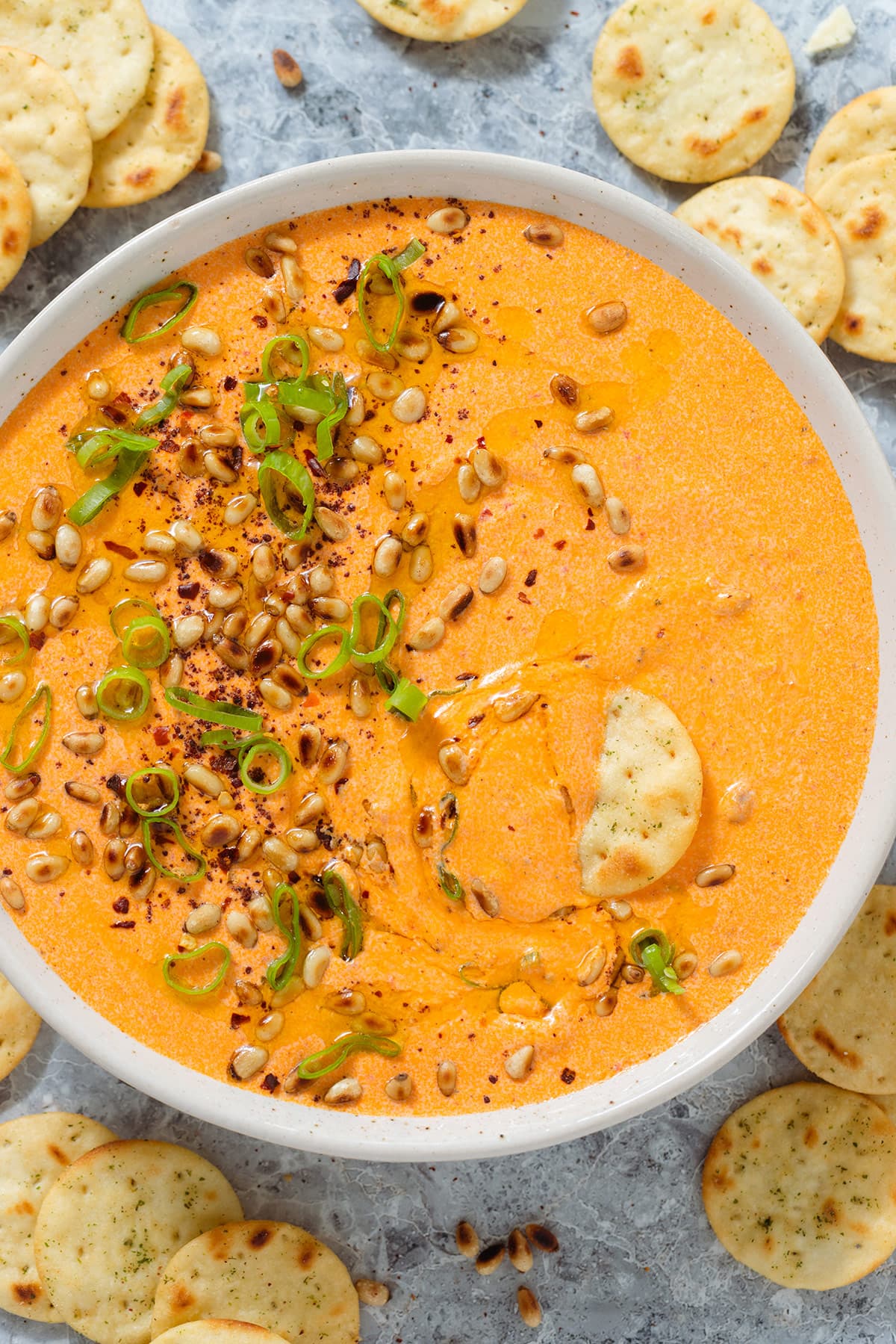 Bright orange dip in a beige bowl garnished with toasted pine nuts, spring onion, and ground sumac, with round crackers around the bowl and one inserted into the dip on the right side of the bowl.