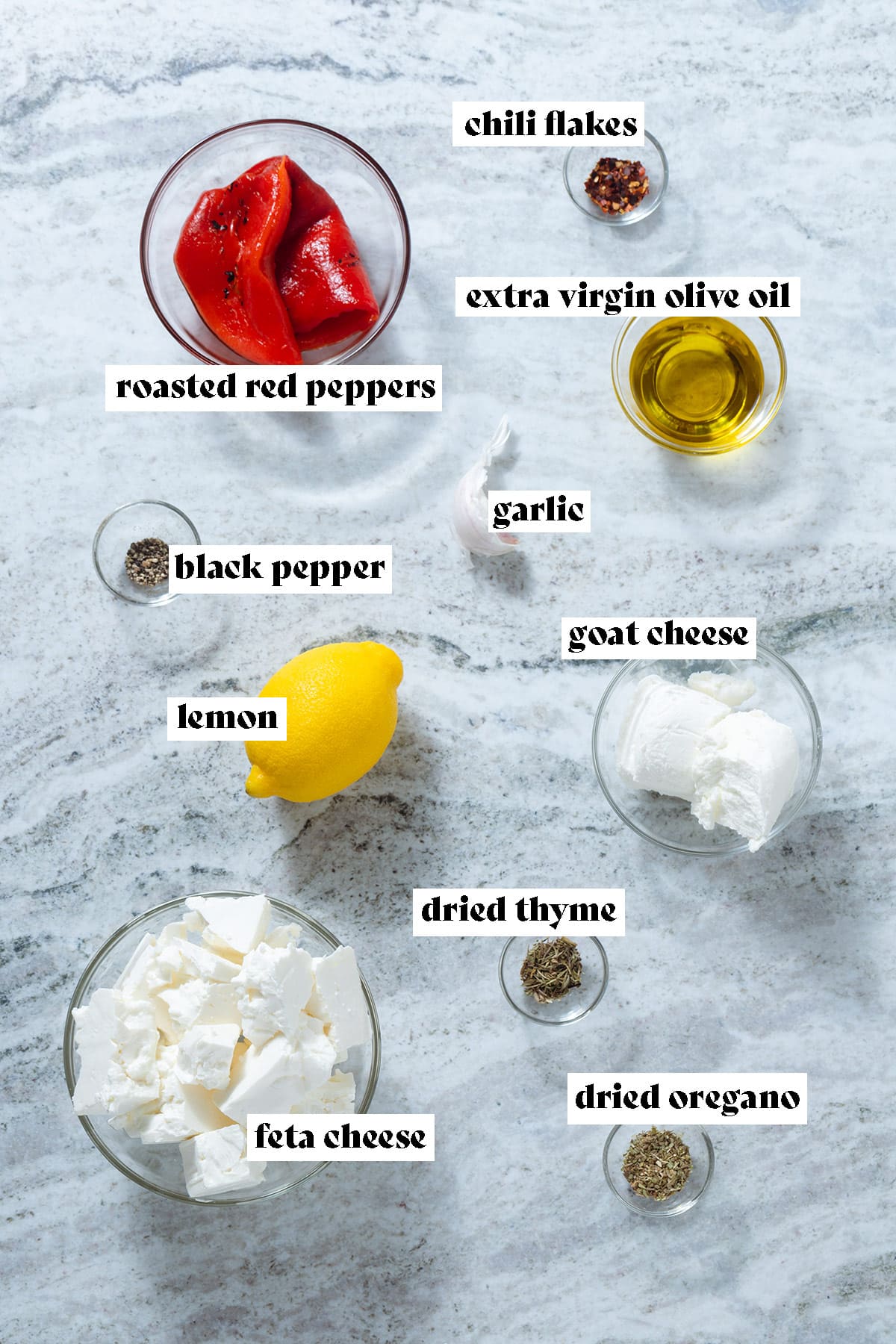 Feta, goat cheese, roasted red pepper, lemon, olive oil, and other ingredients all measured out laid out on a grey stone background with text overlay.