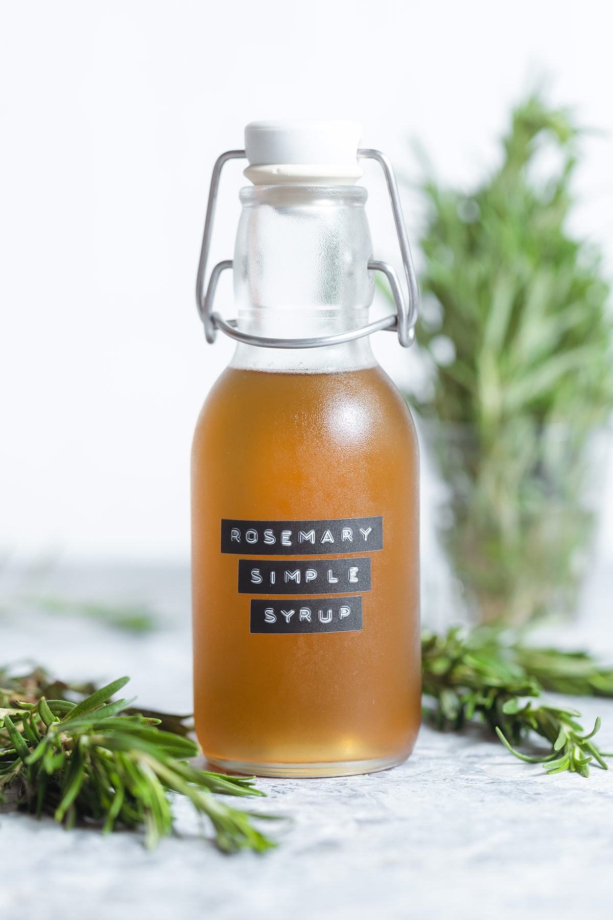 Rosemary simple syrup in a glass jar with more fresh rosemary in the background on a light grey background.