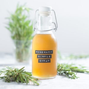 Light yellow rosemary simple syrup in a glass jar with more fresh rosemary in the background on a light grey background.