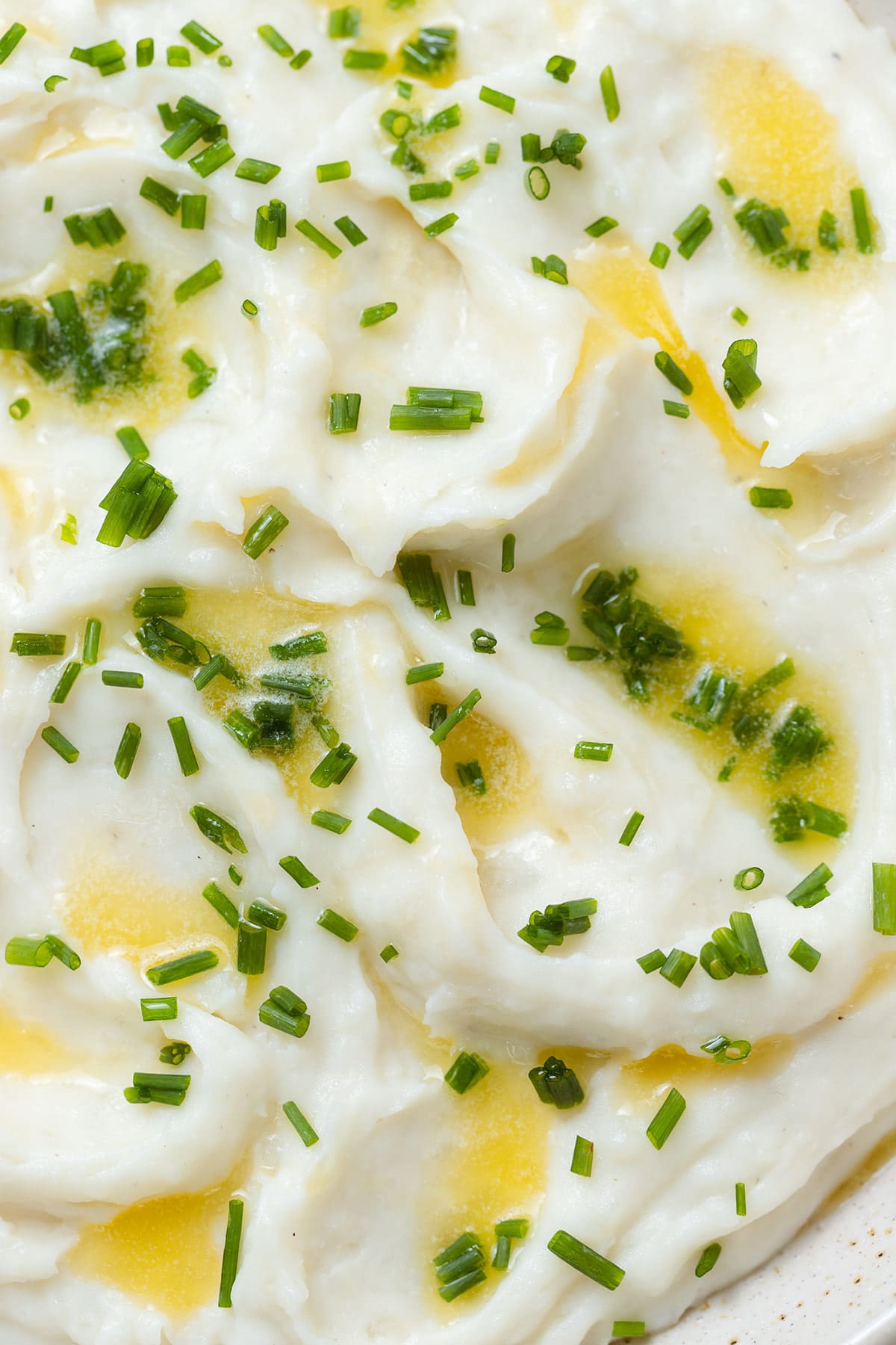 Whipped mashed potatoes topped with melted butter and chopped fresh chives in a beige bowl.
