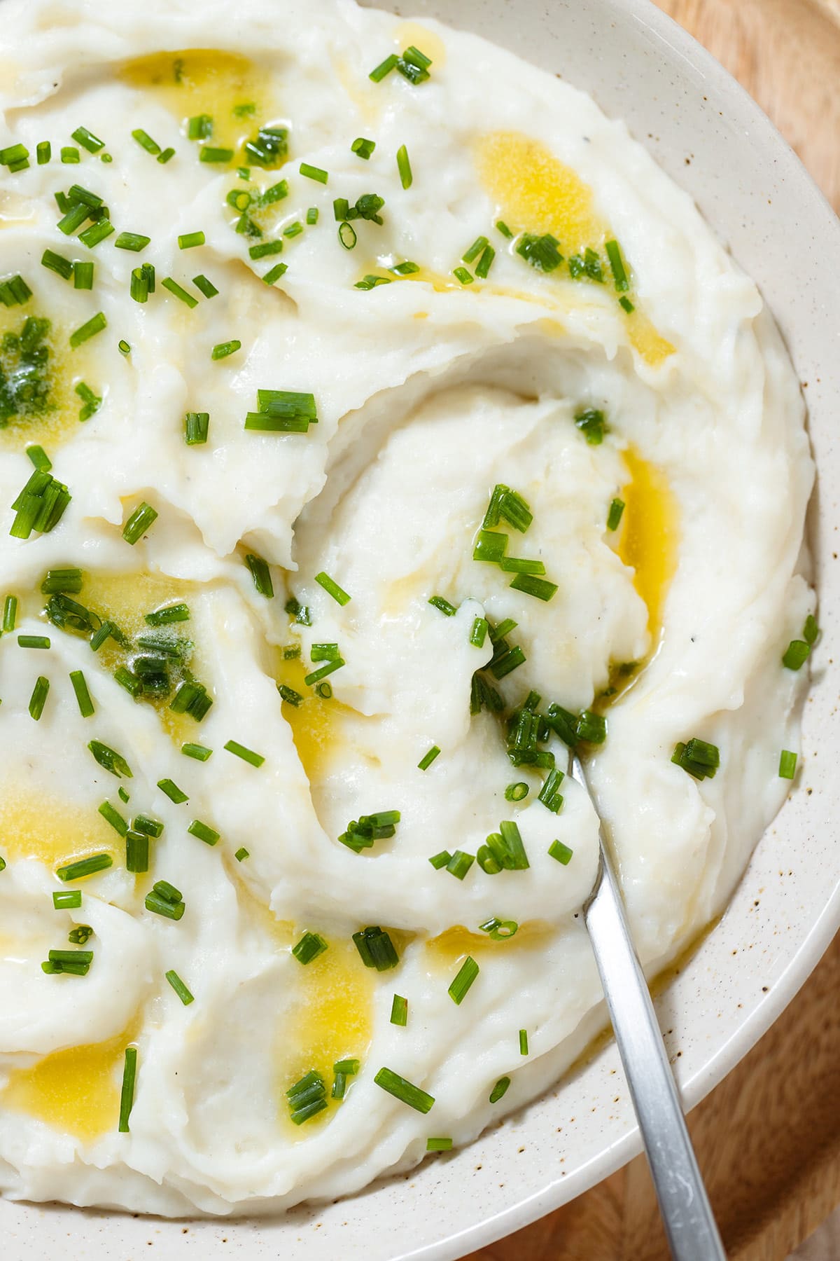 Whipped mashed potatoes topped with melted butter and chopped fresh chives in a beige bowl with a silver spoon inserted on the right.