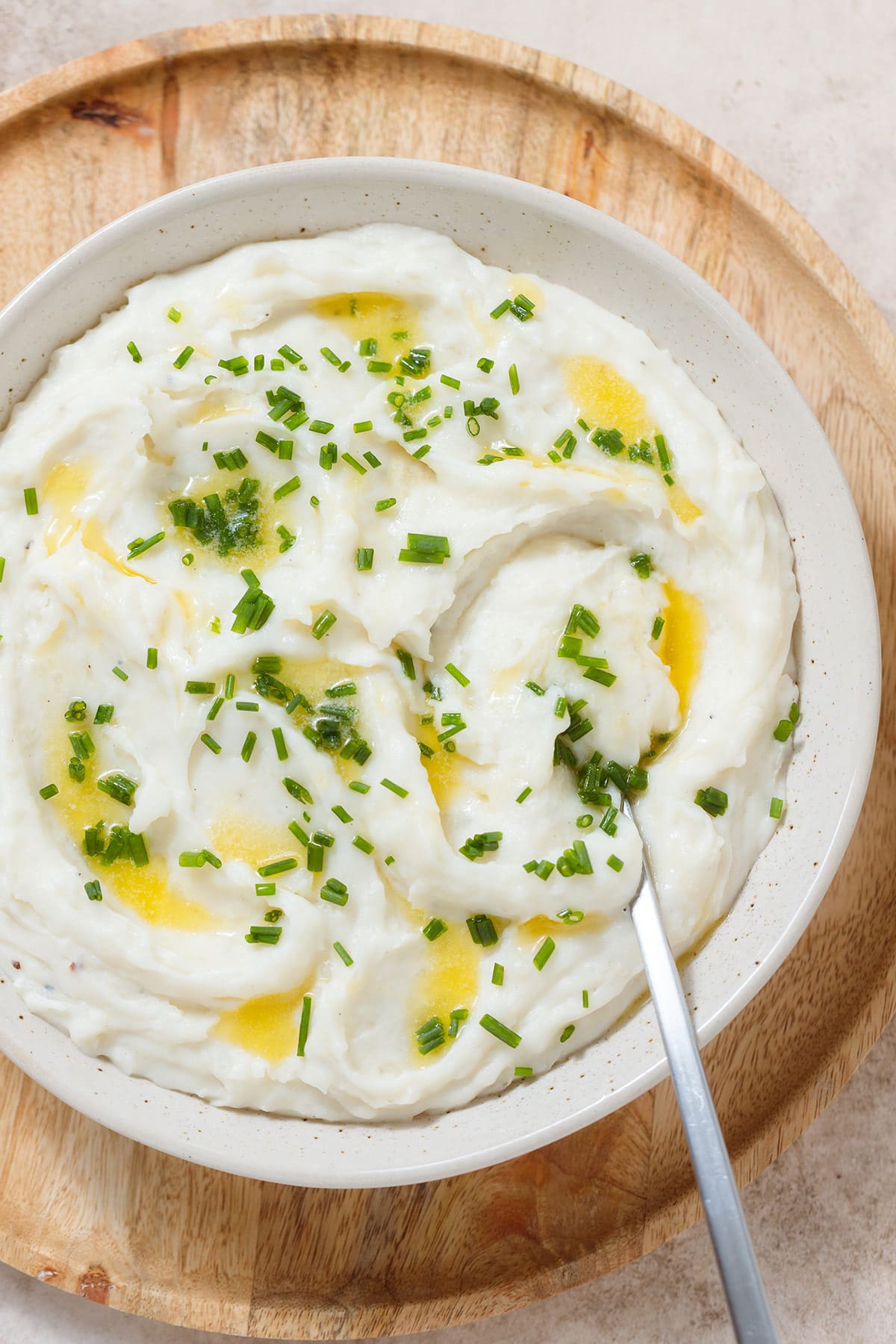 Whipped mashed potatoes topped with melted butter and chopped fresh chives in a beige bowl on a wooden serving plate with a silver spoon inserted on the right.