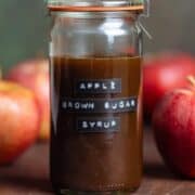 Brown syrup in a glass jar with a label on it that says Apple Brown Sugar Syrup with apples around it on a dark wooden background.