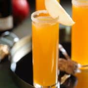 Three tall stemless flute glasses with bright orange apple cider mimosa garnished with a slice of apple and a sugar rim on black background.