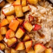 Creamy oatmeal with caramelized apples on top in a beige ceramic bowl with a grey rim on a yellow ceramic plate.