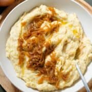 Mashed potatoes topped with caramelized onions in a white bowl with a spoon on the right side on a wooden plate.