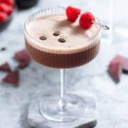 A chocolate espresso martini with rich foam on top in a coupe glass garnished with three coffee beans and a cocktail pick with two raspberries on a grey background.