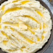 Mashed potatoes in a black low bowl with melted butter on top sprinkled with dried oregano on a grey stone background with a small bottle of truffle oil lying in the top right corner.