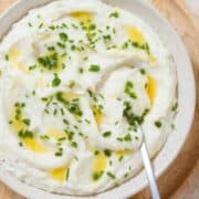 Whipped mashed potatoes topped with melted butter and chopped fresh chives in a beige bowl on a wooden serving plate with a silver spoon inserted on the right.