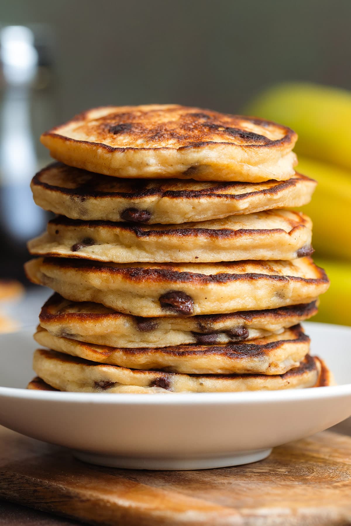 A stack of chocolate chip pancakes on a deep small white plate with bananas in the background.