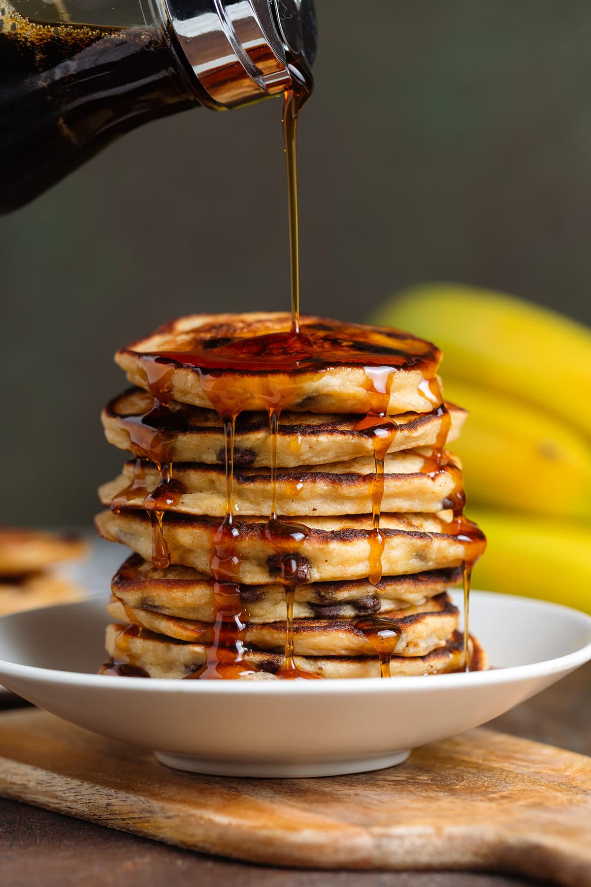 A stack of chocolate chip pancakes on a deep small white plate being drizzled with maple syrup.