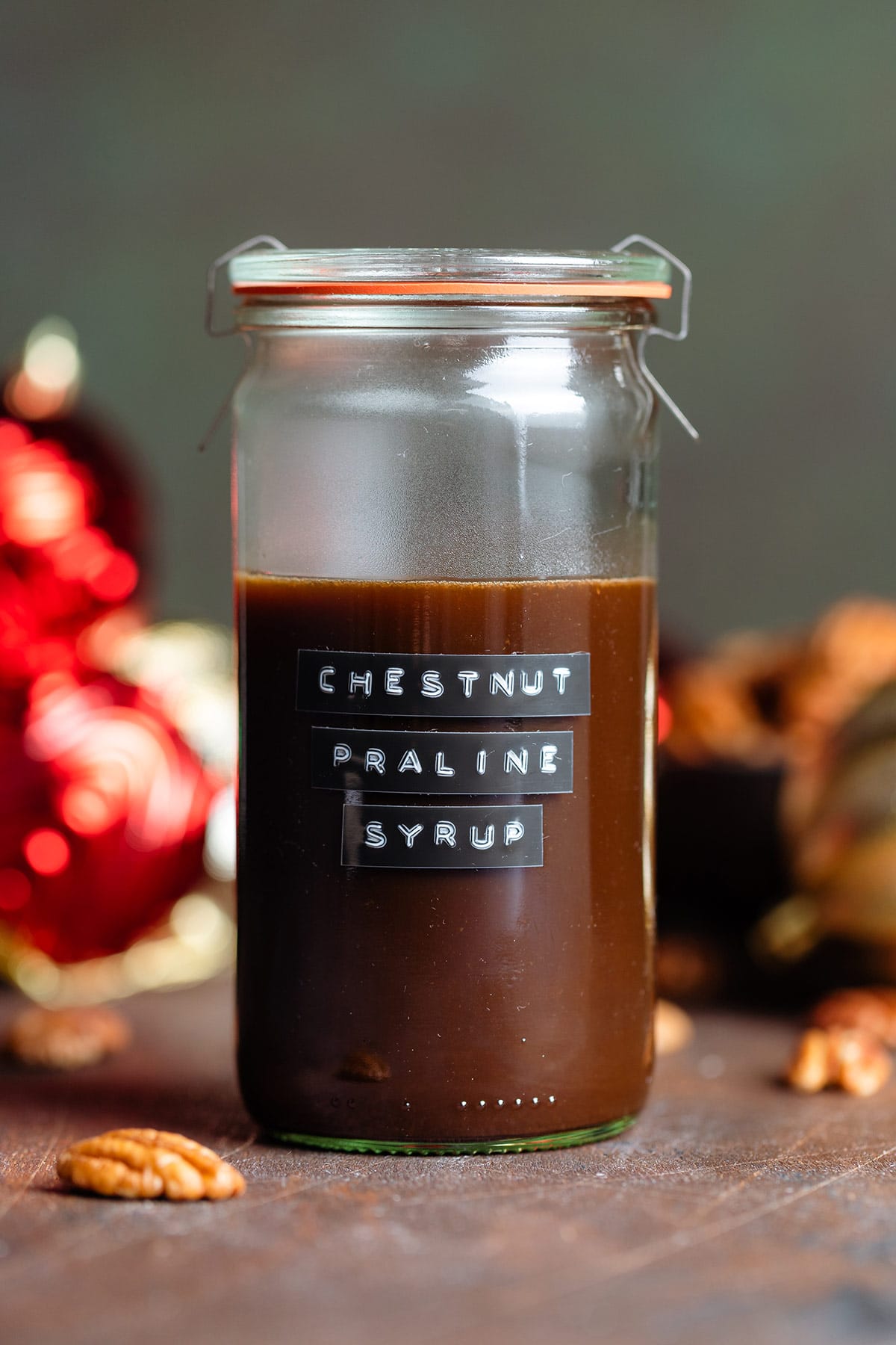 Dark brown chestnut praline syrup in a glass jar with a glass lid and Christmas ornaments in the background.