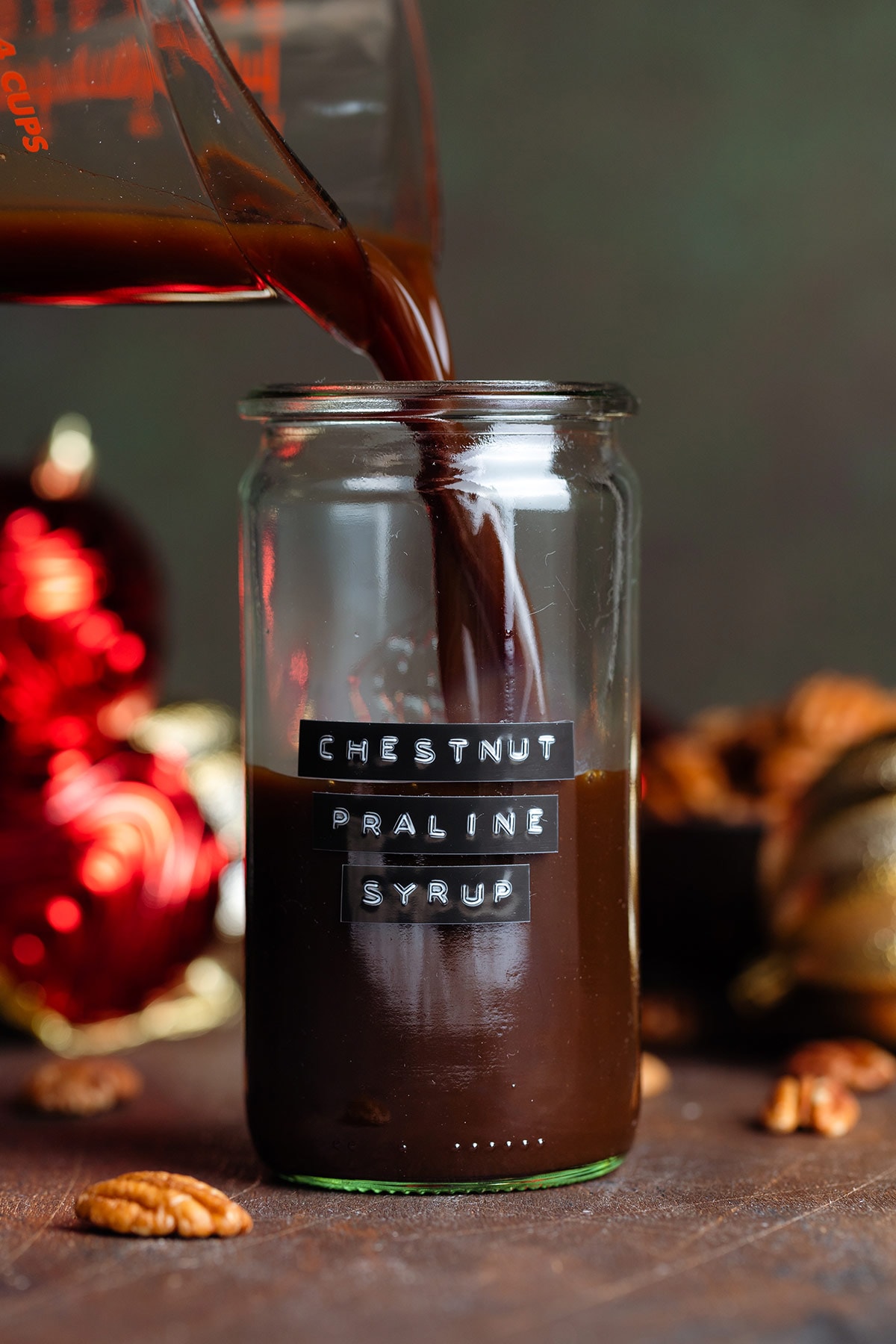 Dark brown syrup being poured into a glass jar with a black label that says chestnut praline syrup with christmas ornaments in the background.