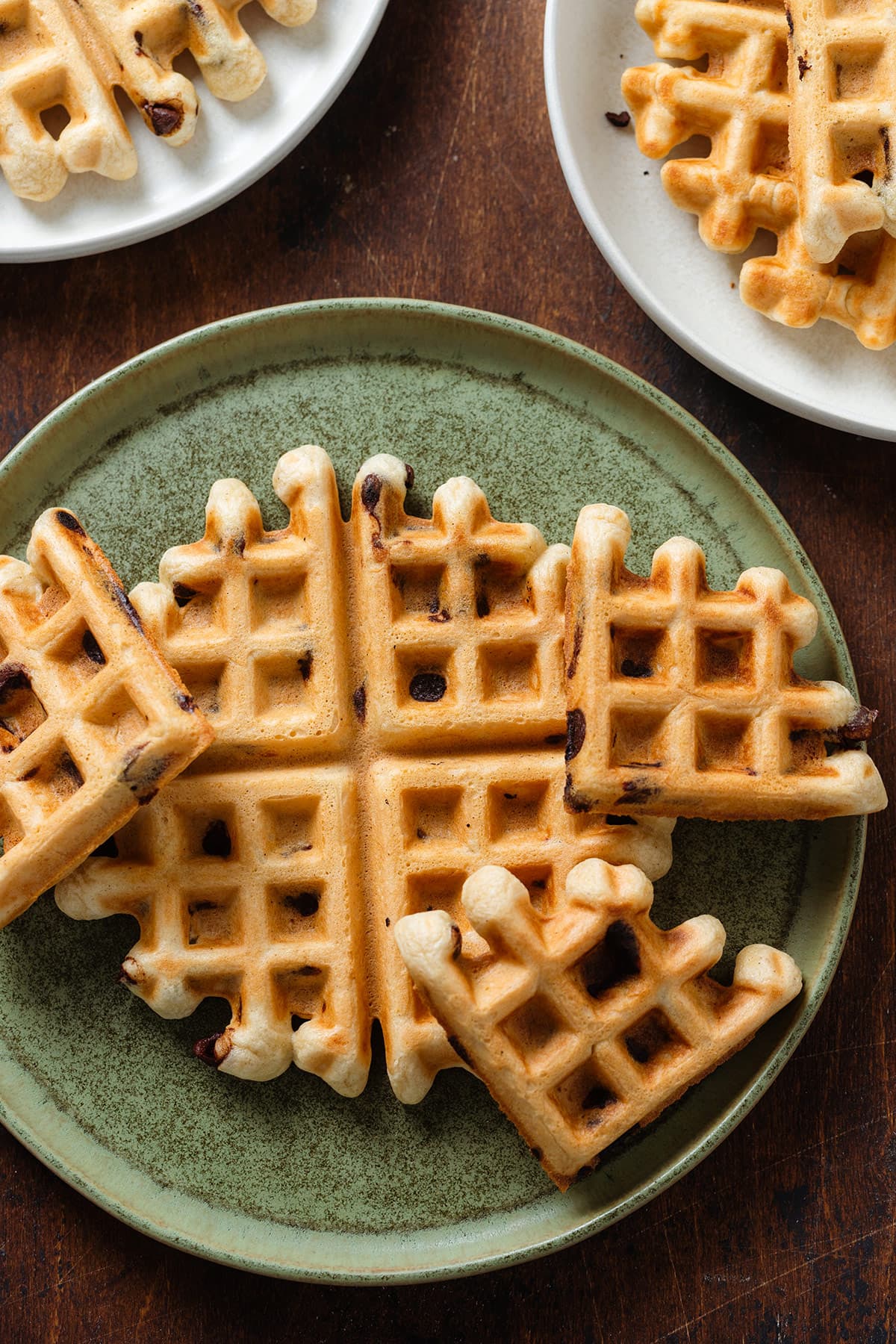 Chocolate chip waffles on a large green plate with more waffles on white plates around it on a dark wooden background.