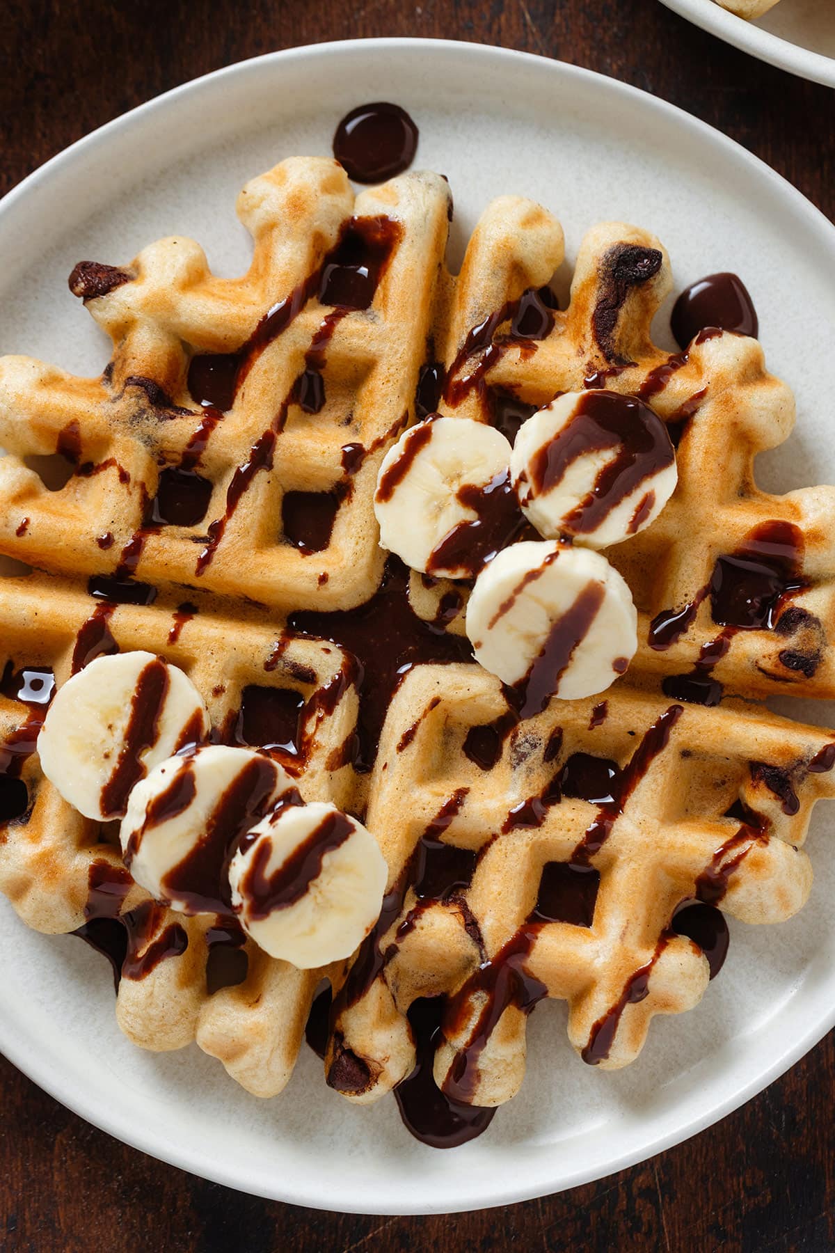 A round waffle on a white plate drizzled with chocolate sauce and maple syrup garnished with sliced banana on a wooden background.