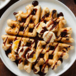 A round waffle on a white plate drizzled with chocolate sauce and maple syrup garnished with sliced banana.