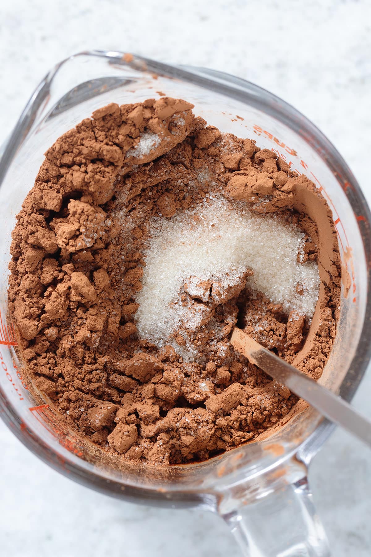 Cane sugar and cacao powder in a large glass measuring cup being mixed together with a spoon.