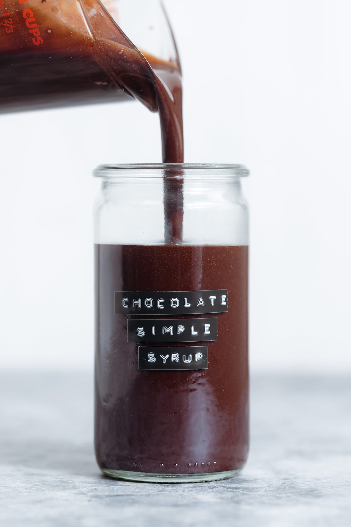 Chocolate syrup being poured into a medium glass jar with a black and white embossed label, on a white background.