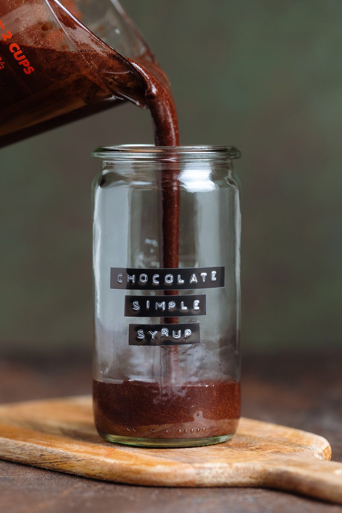 Chocolate syrup being poured into a medium glass jar with a black and white embossed label, on a dark wooden background.