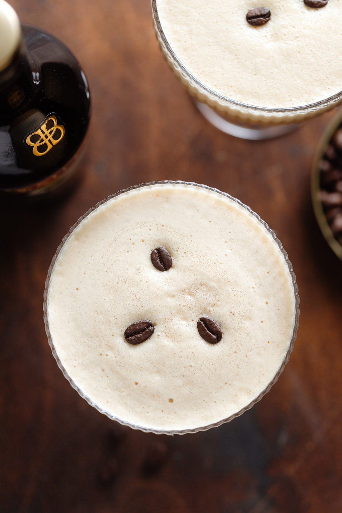 A beige-colored creamy espresso martini with thick foam on top in a coupe martini glass garnished with three coffee beans on a dark wooden background shot from above.