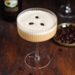 A beige-colored creamy espresso martini in a coupe martini glass garnished with three coffee beans on a dark wooden background.