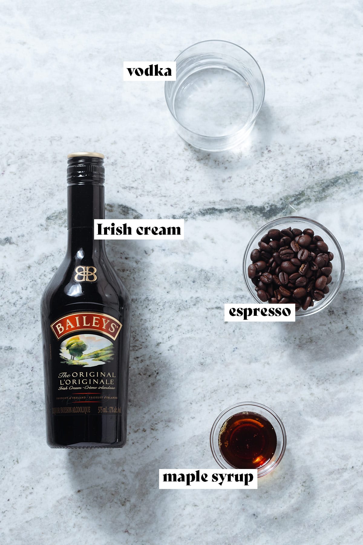 A bottle of Bailey's, maple syrup, vodka, and coffee beans on a grey stone background with text overlay.