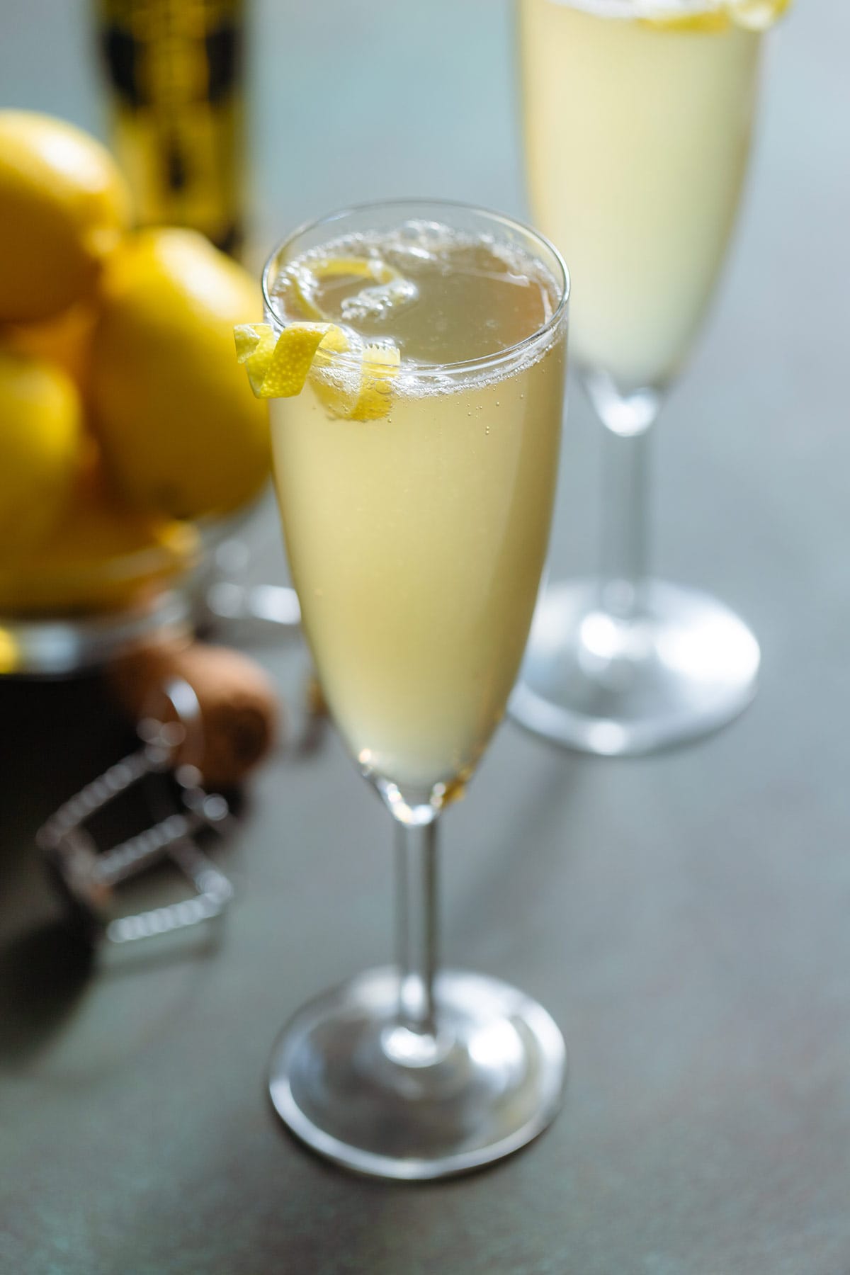 A light yellow French 77 cocktail in a tall champagne flute garnished with a lemon twist on a green background with a bottle of St Germain and a bowl of lemons in the back.