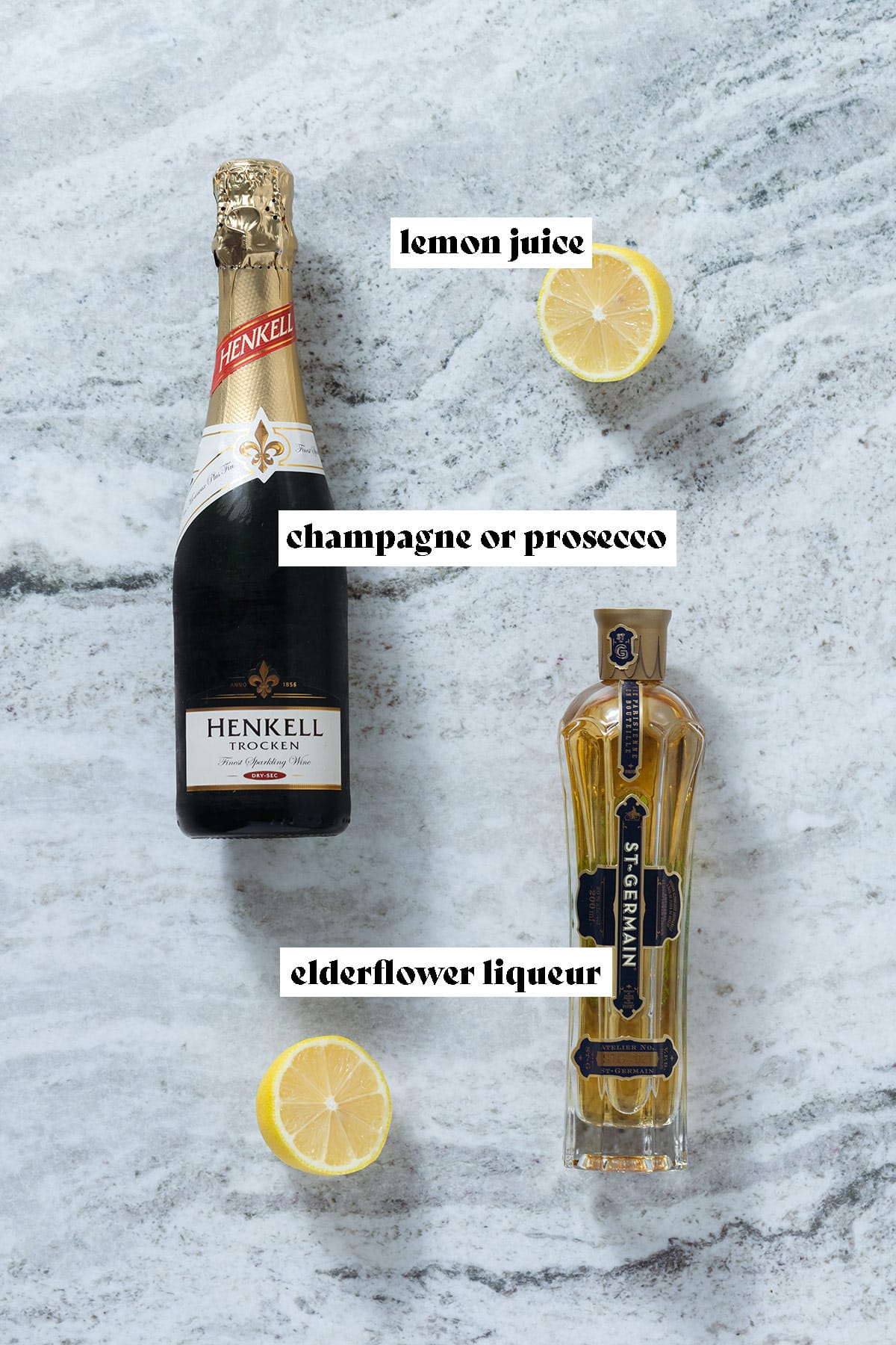 A bottle of Prosecco, St Germain liqueur, and a lemon cut in half on a grey stone background with text overlay.