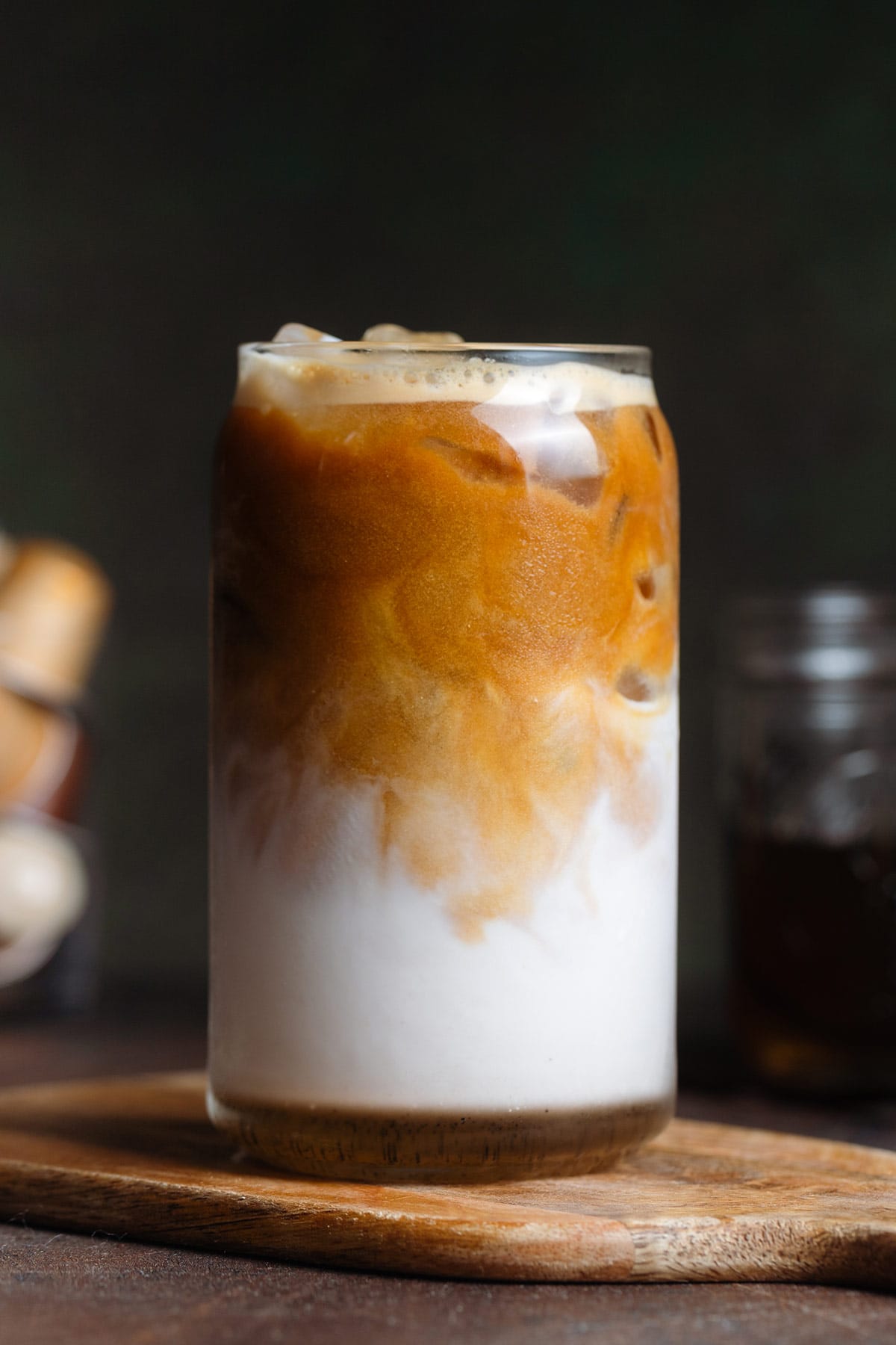 A tall can-shaped glass with iced vanilla latte with the coffee slowly mixing into the milk on a wooden background.