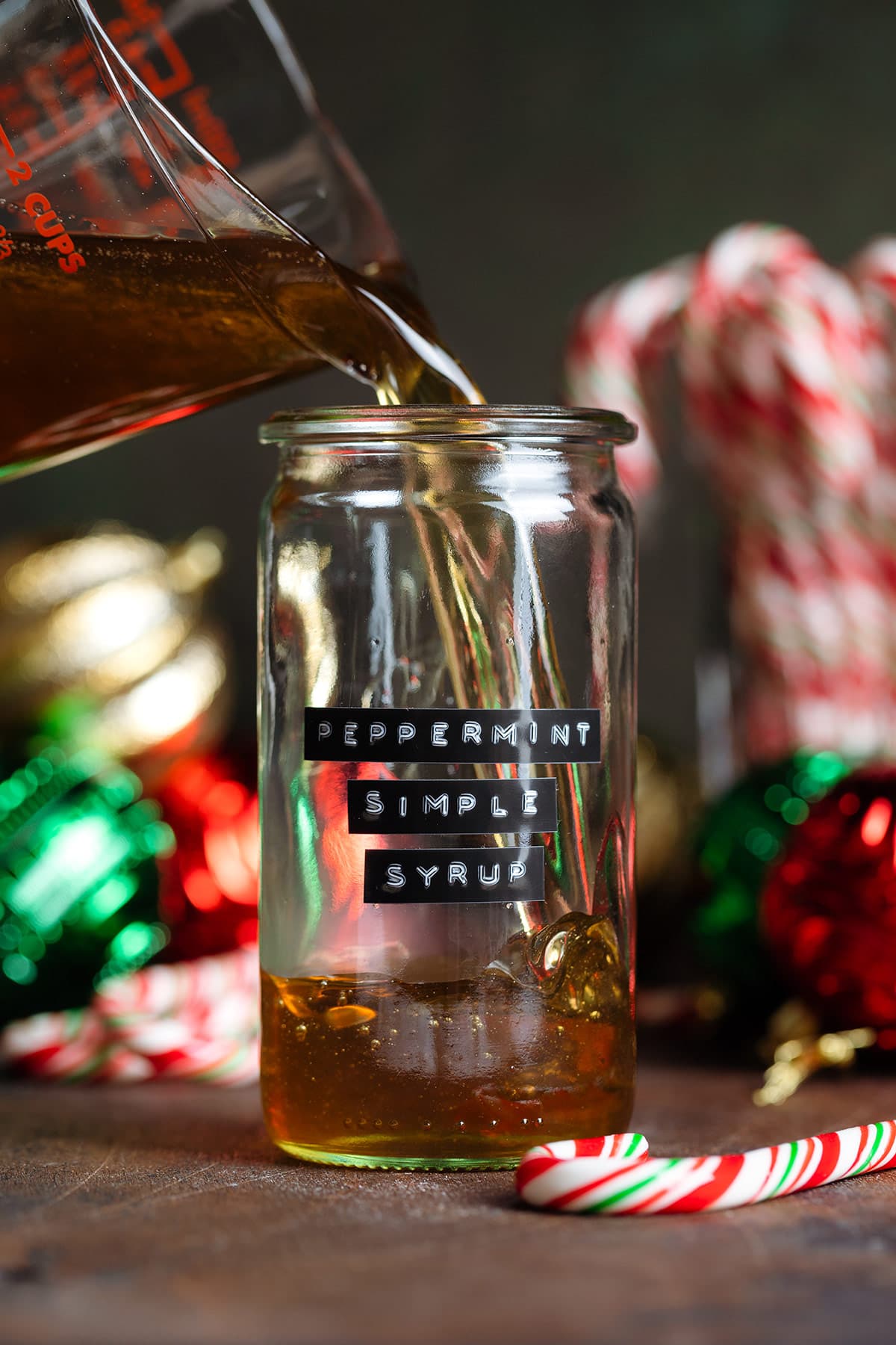 Golden syrup being poured into a glass jar with a black label that says peppermint simple syrup on the side with Christmas ornaments in the background.